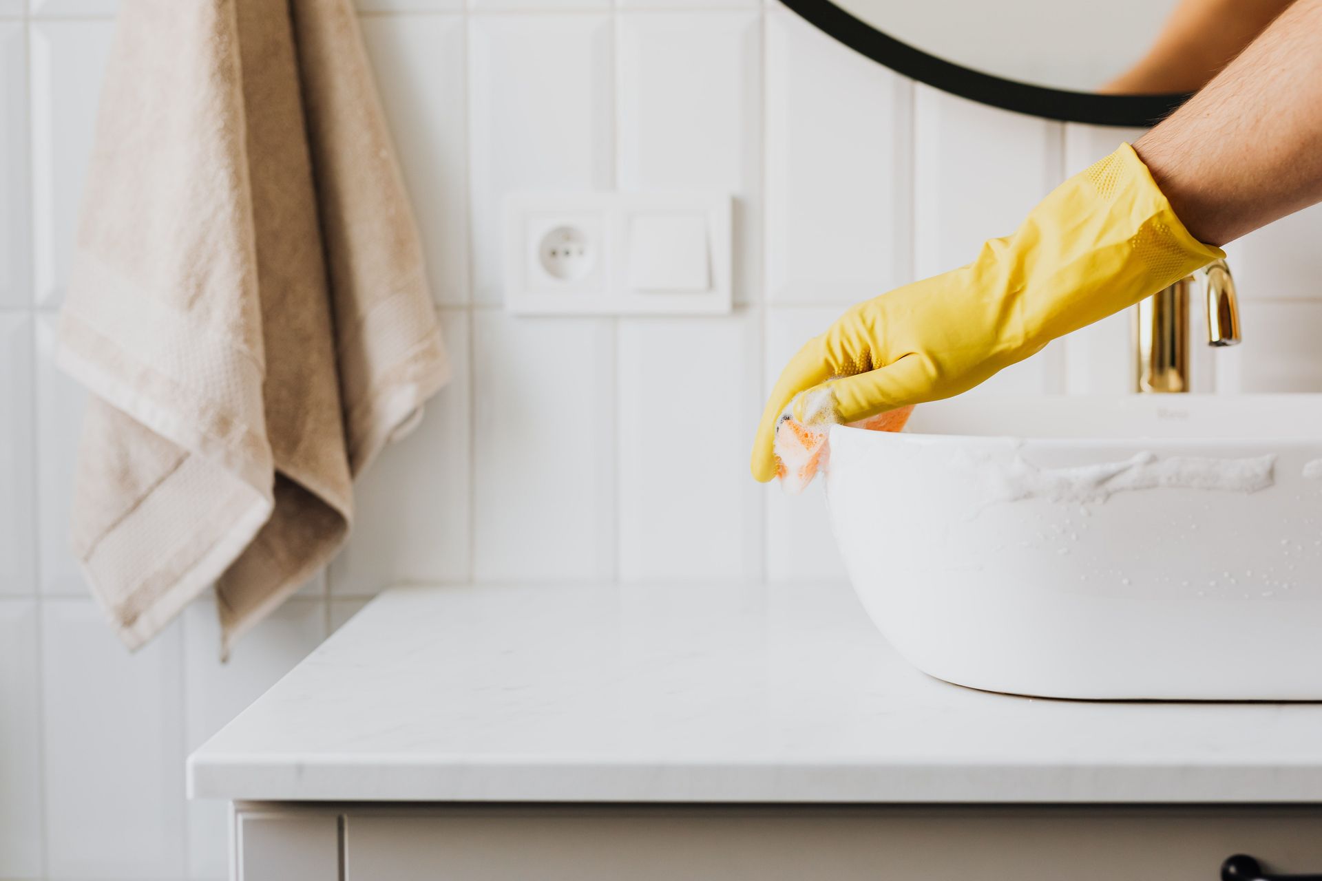 Crop anonymous person wearing yellow latex gloves washing sink in bathroom with orange sponge and cleanser while cleaning contemporary flat